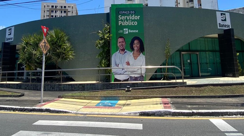 Imagem da fachada da agência do Banese em frente ao Shopping Jardins, com um banner grande na fachada onde se lê "Espaço do Servidor Público, e embaixo desse texto pousam um homem e uma muher negra de braços cruzados, ambos sorriem.