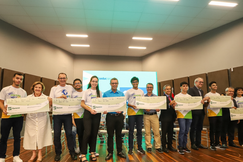 Autoridades entregam prêmio, em formato de cheque grande simbólico, aos estudantes da rede pública estadual com melhores notas no Enem 2023. Entre as autoridades estão o governador Fábio Mitidieri, e o diretor de Tecnologia do Banco, Kleber Teles.