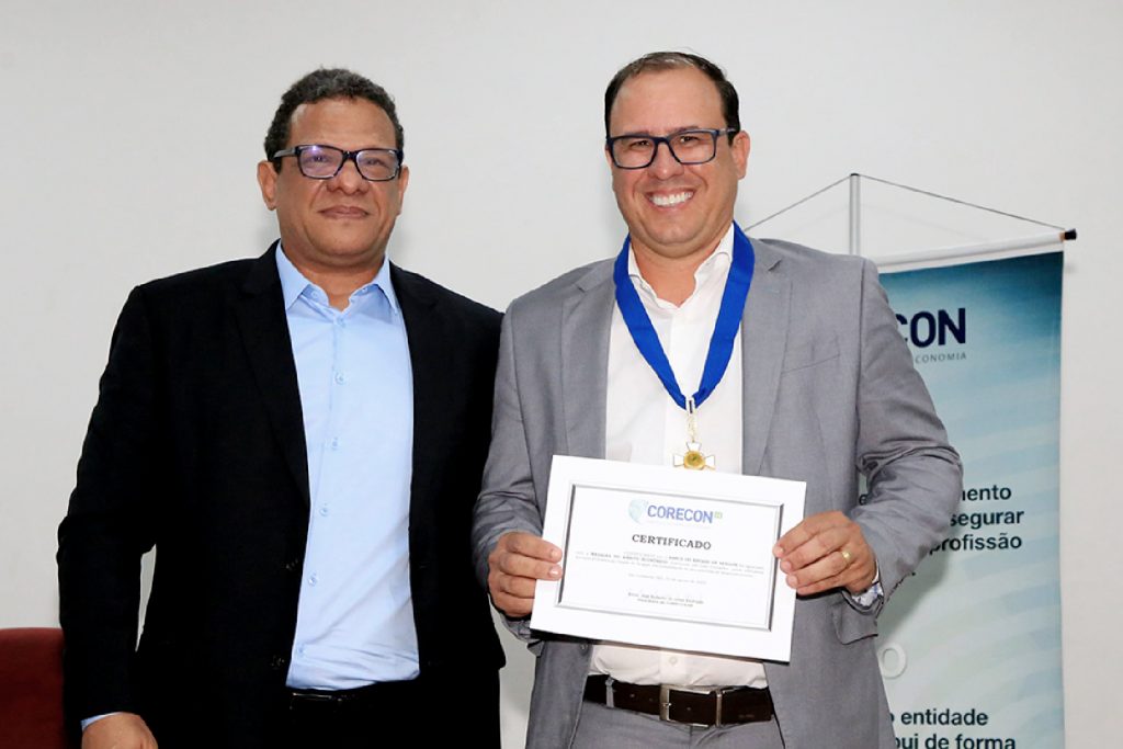 O presidente do Corecon-SE, José Roberto de Lima Andrade, ao lado do diretor de Crédito e Serviços do Banese, Wesley Cabral, que mostra o certificado da condecoração e posa com a Medalha de Mérito pendurando no pescoço em cordão azul. 